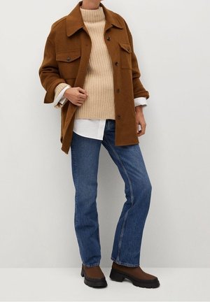 Personne portant une veste marron à boutons sur un pull côtelé beige, une chemise blanche, un jean bleu et des bottes marron, debout contre un mur blanc.