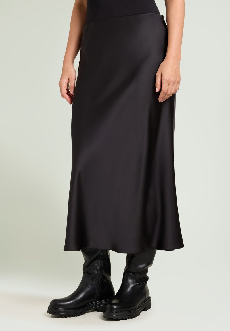 Schwarze Satin-Maxi-Rock mit glatter Textur, leichtem Glanz und ausgestelltem Saum, kombiniert mit klobigen schwarzen Lederstiefeln bis zum Knie.
