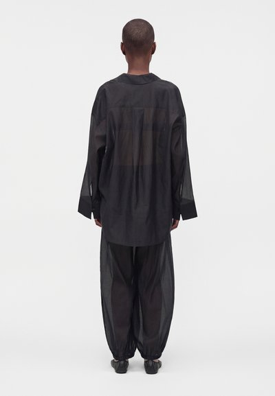 Personne debout, de dos, portant une chemise noire oversize transparente et un pantalon ample assorti avec des chevilles resserrées et des chaussures noires.