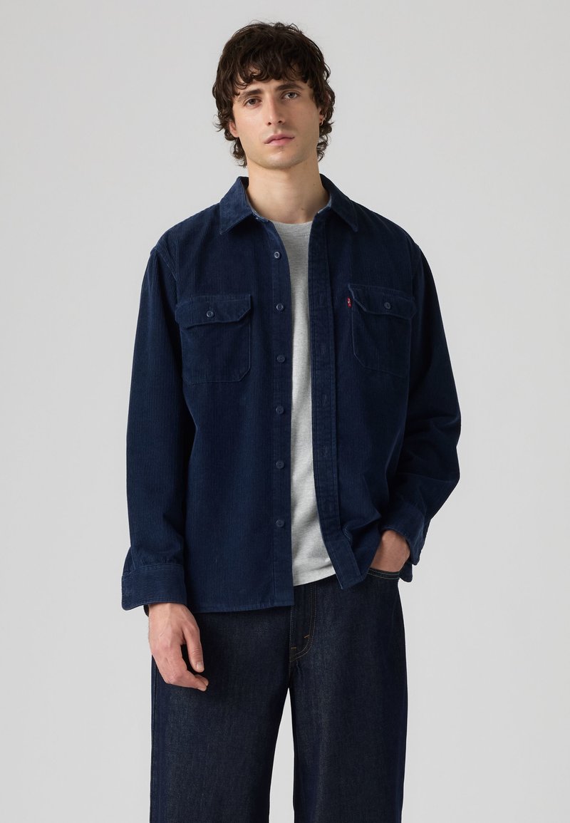 Homme portant une chemise en velours côtelé bleu foncé par-dessus un t-shirt gris clair et un jean bleu foncé, debout avec une main dans la poche sur un fond uni.