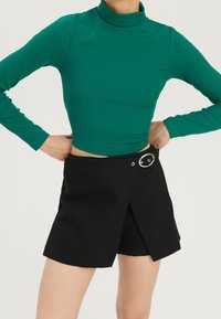 Schwarze Wickelhosen mit silbernem Akzent und Knopfverschluss, kombiniert mit einem enganliegenden, gerippten, langärmligen Rollkragenoberteil in leuchtendem Grün.