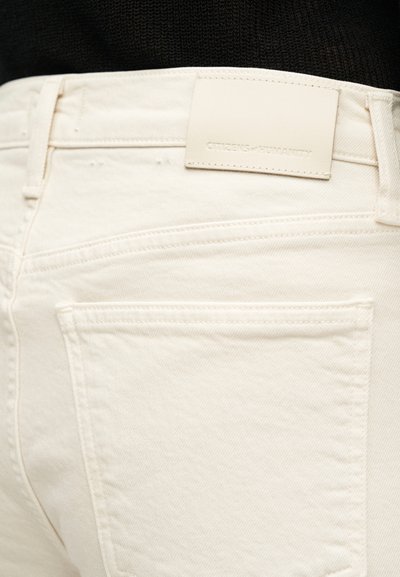 Κοντινό πλάνο της πίσω τσέπης και της μέσης λευκού τζιν, με ραμμένο ανοιχτόχρωμο δερμάτινο έμβλημα που γράφει "CITIZENS of HUMANITY."