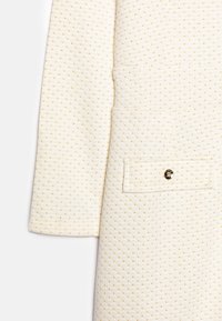 Weißer strukturierter Stoffmantel mit kleinen goldenen Punkten, einer einzelnen Tasche mit Knopf und einer klaren, strukturierten Silhouette.