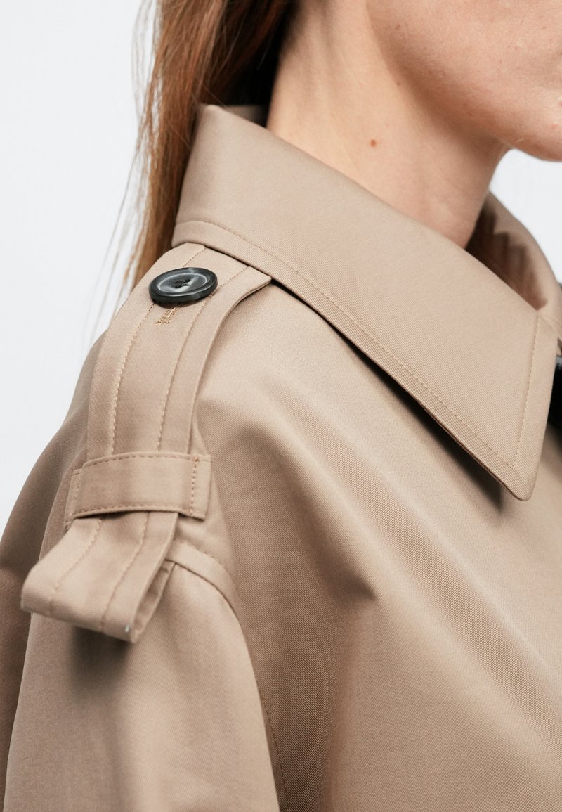 Trench-coat beige avec un large col, des épaulettes et un détail de bouton noir. Texture lisse avec une silhouette structurée.