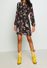 Schwarz mit floralen Mustern, langärmliges Kleid mit Schlüsselloch-Ausschnitt; kombiniert mit leuchtend gelben Cowboy-Stiefeln mit weißen Akzenten.