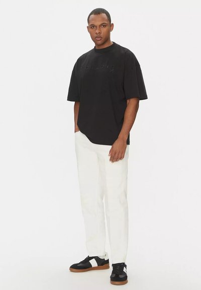 Uomo in piedi che indossa una t-shirt nera di taglia oversize, pantaloni bianchi e sneakers bianco e nere, con uno sfondo chiaro e semplice.