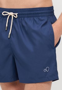 Marineblaue Badeshorts aus leichtem Stoff; elastischer Bund mit beigem Kordelzug; Seitentaschen; kleines weißes Logodetail.