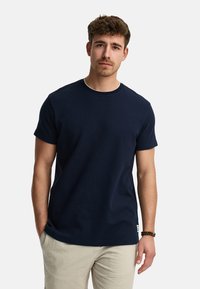 Camiseta de algodón azul marino con mangas cortas y cuello redondo, que presenta una textura sutil y una pequeña etiqueta de logo en el dobladillo inferior.