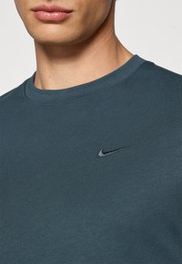 Tume teali värvi puuvillane T-särk ümmarguse kaelusega ja lühikeste varrukatega. Rinnal on väike must Nike logo tikandina.