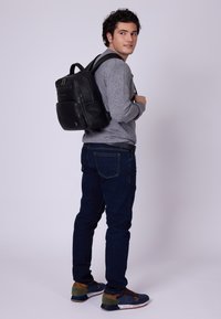 Ung man i grå tröja, mörka jeans och blå sneakers, bärande på en svart ryggsäck, tittar över sin axel mot en neutral bakgrund.