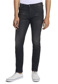 Person som bär slim-fit svarta jeans och vita sneakers, står mot en vit bakgrund med händerna avslappnat vid sidorna.