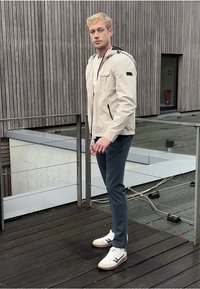 Ljust beige zip-up jacka med huva, två sidofickor, som bärs med mörka byxor och vita sneakers med svarta accenter. Träbakgrund.