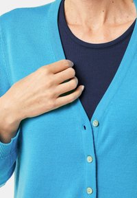 Hellblauer Cardigan aus Strickstoff, mit V-Ausschnitt, fünf Knöpfen und gerippten Bündchen. Getragen über einem dunkelblauen Oberteil.