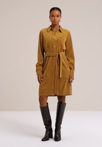 Abito camicia in corduroy senape con maniche lunghe, chiusura a bottoni e cintura. Abbinato a stivali marroni fino al ginocchio per un contrasto.