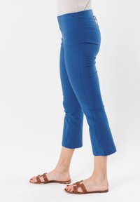 Pantaloni capri blu in tessuto liscio, design aderente, vita media e orli rifiniti. Indossati con sandali a infradito marroni.