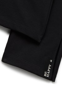 Czarna tkanina z bliska, pokazująca brzeg z wyszywaną metką z napisem "BĄDŹ SZCZĘŚLIWY" w białym kolorze oraz małym symbolem powyżej.