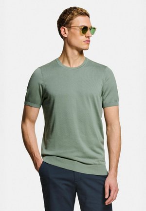 Homme portant un pull léger vert clair à manches courtes et un pantalon bleu marine foncé, avec des lunettes de soleil à monture dorée et verres verts, posant avec une main dans la poche.