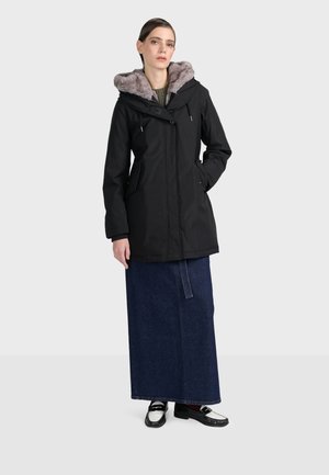 Parka noire avec capuche en fausse fourrure, dotée d'une fermeture éclair et de boutons, de deux poches à l'avant et d'une coupe décontractée. Portée avec une longue jupe en denim.