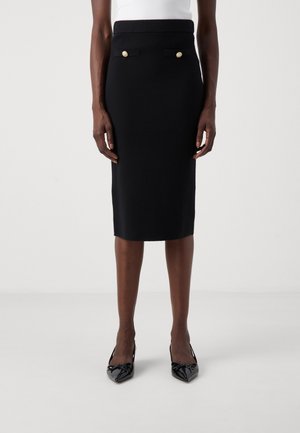 Modelka nosząca wysoką, do kolan, czarną ołówkową spódnicę z detalami w postaci złotych guzików oraz czarne szpilki z noskiem w szpic.