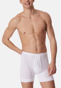 Vita bomullsboxer med ribbad textur, elastisk midja med varumärkeslogotyp och en öppning framtill. Modellen står och ler.