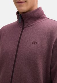 Felpa zip-up bordò con collo alto, caratterizzata da una superficie texturizzata e un piccolo logo sul lato sinistro del petto. La cerniera è in metallo nero.