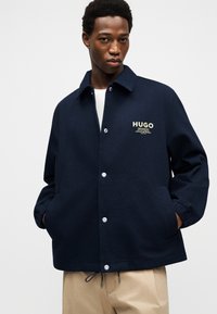 Ung mand iført en marineblå jakke med knapper og "HUGO"-logo på brystet samt beige bukser, stående med hænderne i lommerne.