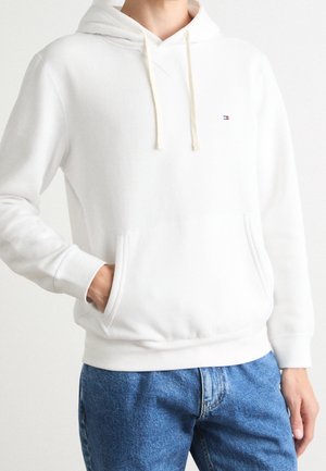 Person trägt ein weißes Kapuzen-Sweatshirt mit Fronttaschen und kleinem Logo auf der Brust, kombiniert mit blauen Jeans vor einem schlichten Hintergrund.