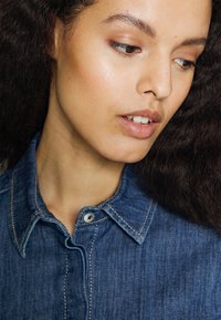 Camisa de ganga com colarinho pontudo, fecho de botões e costura visível. O tecido é azul médio com uma leve textura.