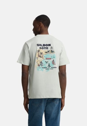 Hombre vistiendo camiseta blanca con ilustración colorida de la playa de la Riviera Francesa y texto en la espalda, combinada con jeans azules.