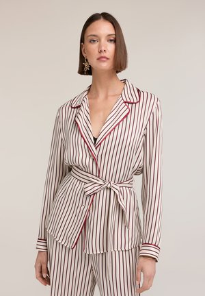 Donna con taglio bob liscio che indossa un completo stile pigiama bianco con righe rosse verticali, cintura annodata in vita e orecchino dorato e vistoso.