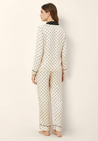 Polka dot pajama set in cream with black dots, long sleeves, and contrasting black collar and cuffs. Smooth fabric, relaxed fit, ankle-length pants.