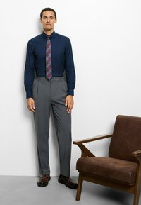 Dunkelblaues Hemd mit gestreifter Krawatte, graue Hose und braune Schuhe. Nebenan ein brauner Sessel. Einfaches Design, glatte Oberfläche.