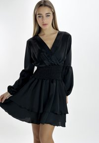 Zwarte satijnen jurk met een diepe V-hals, gesmockte tailleband, lange mouwen en een gelaagde rok. Transparante stof benadrukt een vloeiende silhouet.