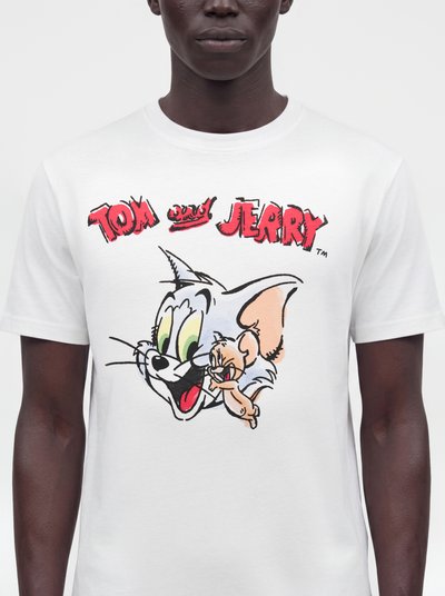 T-shirt blanc avec texte rouge « Tom and Jerry » et visages de dessins animés de Tom le chat et Jerry la souris au centre, porté par une personne debout.