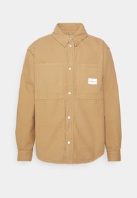 Camicia in cotone marrone con colletto, maniche lunghe e due tasche frontali. Presenta bottoni a pressione e un'etichetta del marchio sulla tasca.