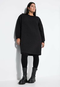 Vestido negro de gran tamaño con textura y mangas largas, combinado con leggings negros y botas negras contundentes, todo sobre un fondo claro.