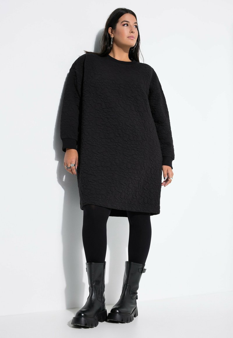 Vestido negro de gran tamaño con textura y mangas largas, combinado con leggings negros y botas negras contundentes, todo sobre un fondo claro.