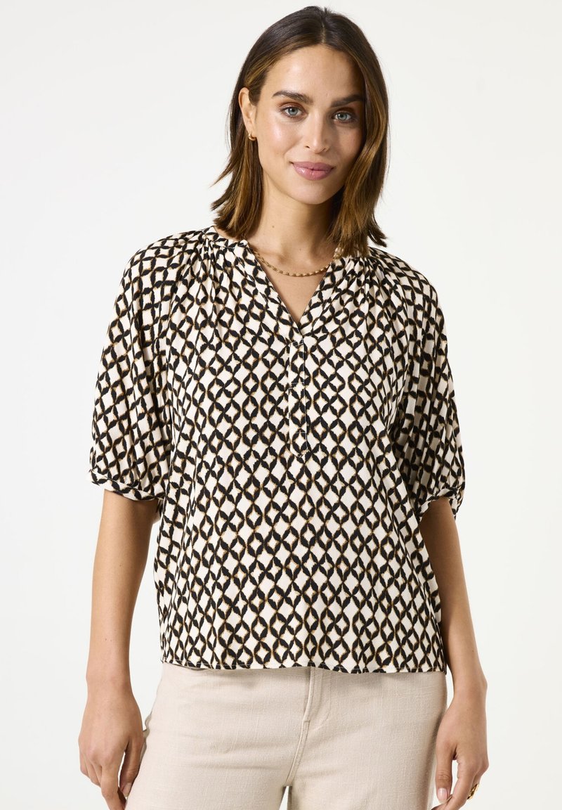 Blouse à manches courtes en tissu crème avec un motif géométrique noir, présentant un décolleté en V et des détails froncés aux épaules.