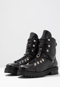 Bottines en cuir noir à lacets avec une tige haute, quincaillerie argentée, semelles robustes et finition texturée. Présence de crochets et d'œillets le long de l'arbre.
