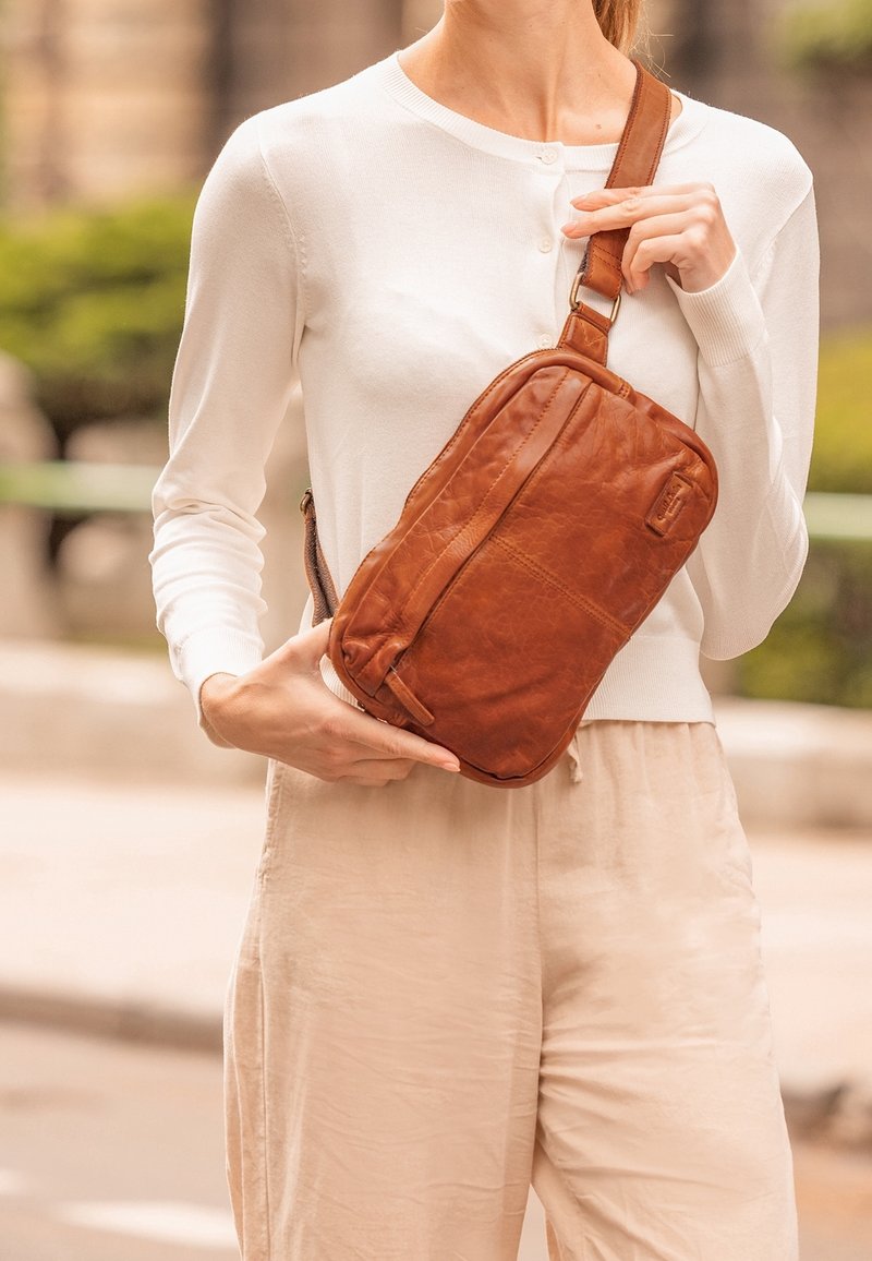Sac en cuir marron en bandoulière avec une texture lisse, comportant une poche zippée et une sangle réglable. Exposé dans un cadre extérieur décontracté.