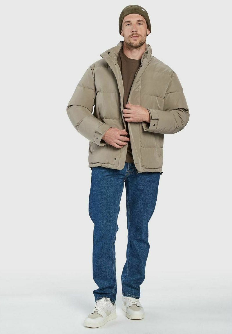 Mann in einer khakifarbenen Steppjacke, braunem Beanie, braunem Hemd, blauen Jeans und weißen Sneakers, der den Reißverschluss seiner Jacke schließt, während er steht.