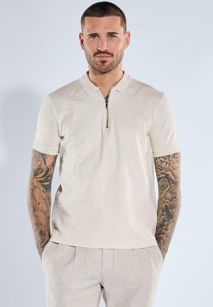 Man with short hair and full sleeve tattoos wears a beige short-sleeve zip-up polo shirt and light-colored pants, hands in pockets.