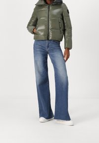 Giacca a piumino imbottita verde con collo alto, chiusura con cerniera e dettagli delle tasche, abbinata a jeans blu a gamba larga e sneakers bianche.