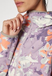 Chaqueta con estampado floral en tonos púrpura y durazno, con cierre de cremallera, diseño ajustado y tejido texturizado. Presenta una capucha y ribetes en contraste.