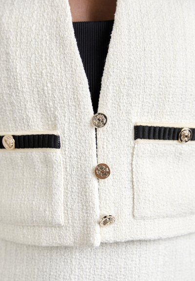 Weißer strukturierter Blazer mit goldenen Knöpfen und schwarzen Taschenakzenten, im taillierten Schnitt und mit V-Ausschnitt.
