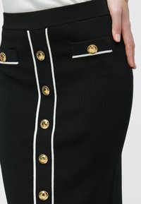 Μαύρη pencil φούστα με λευκή περικοπή, διακοσμημένη με κουμπιά σε χρυσό τόνο και δύο μπροστινές τσέπες, κατασκευασμένη από υφασμάτινη υφή.