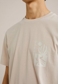 Camiseta de algodón de color beige claro con mangas cortas, que presenta un sutil diseño floral y la palabra "PARADI" en una impresión rectangular.