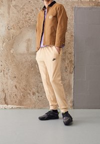 Ruskea puuvillatakk, violetti paita, beige fleecepipot housut mustalla Nike-logolla ja mustat urheilukengät minimalistisessa ympäristössä.