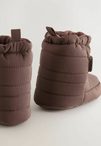 Braune gepolsterte Hausschuhe mit einem gesteppten Design, ausgestattet mit einem weichen, strukturierten Innenfutter, einem verstellbaren Riemen und einem Ziehgriff für ein einfaches An- und Ausziehen.