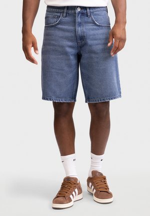 Mężczyzna ubrany w średnio wyprane szorty jeansowe, białe skarpetki do kostek i brązowe zamszowe trampki z białymi paskami na jednolitym tle.